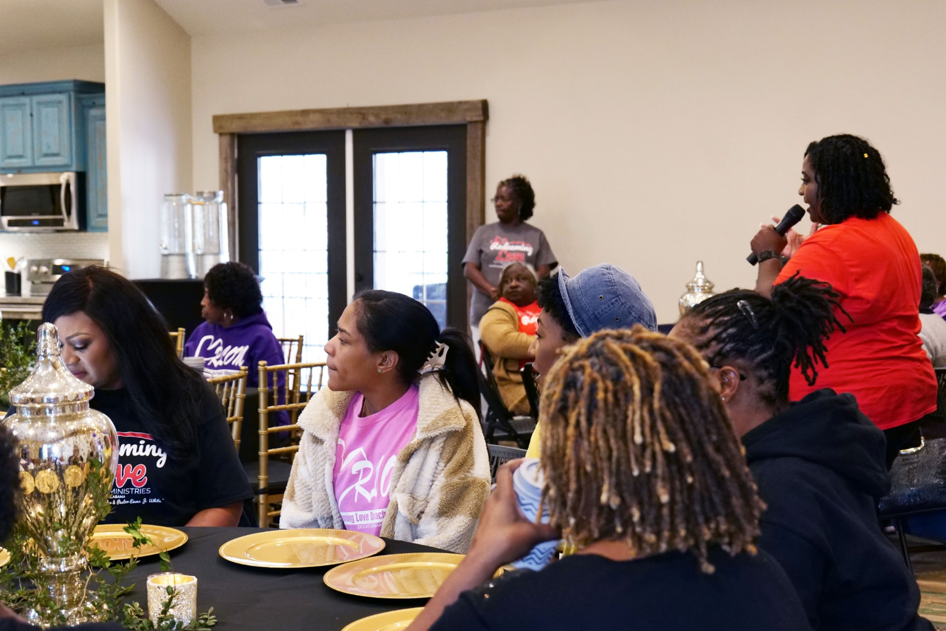 Rose Sherrod speaking into a microphone to women seated at gathering tables during a Daughters of Promise event.