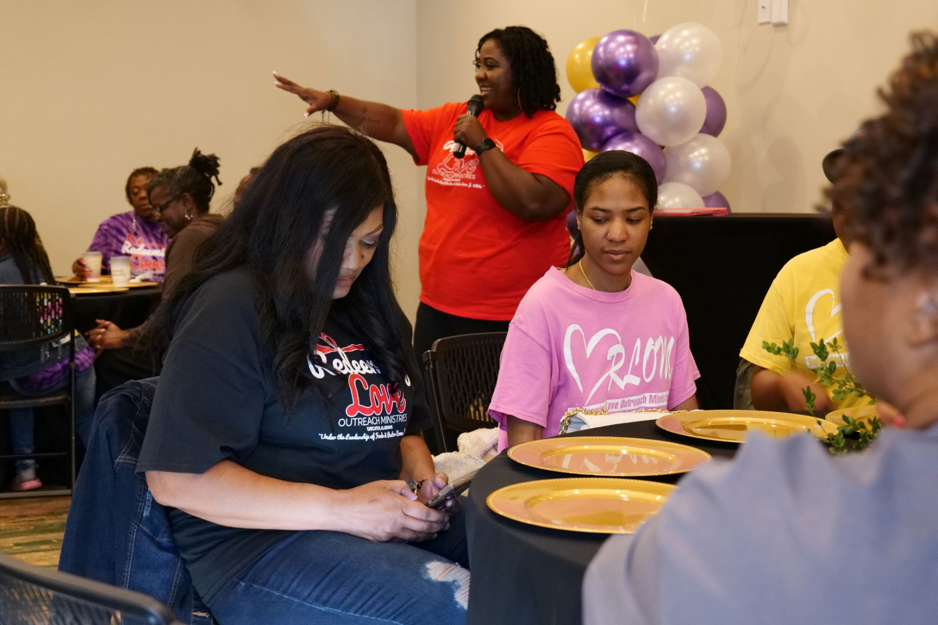 A ministry leader teaching with a microphone while women listen at a Redeeming Love Outreach Ministries event.