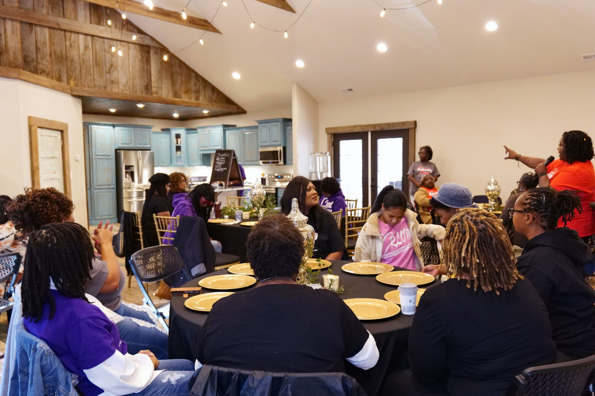 Wide view of a women's ministry gathering, with attendees seated around round tables under string lights.